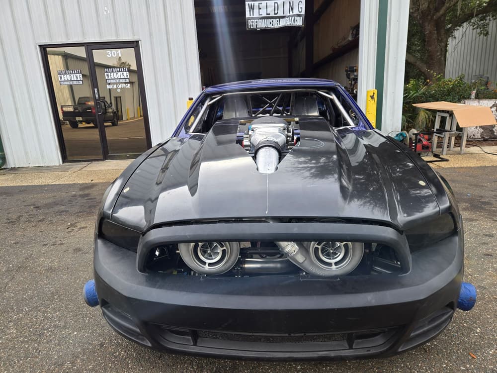 Black twin-turbo drag Mustang parked in front of the Performance Welding shop.